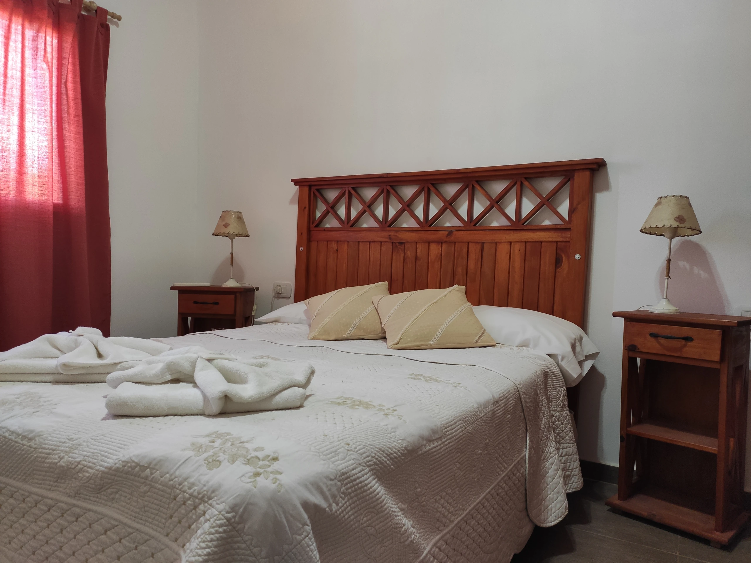 Cama matrimonial con respaldo de madera, mesas de luz y cortinas rojas.