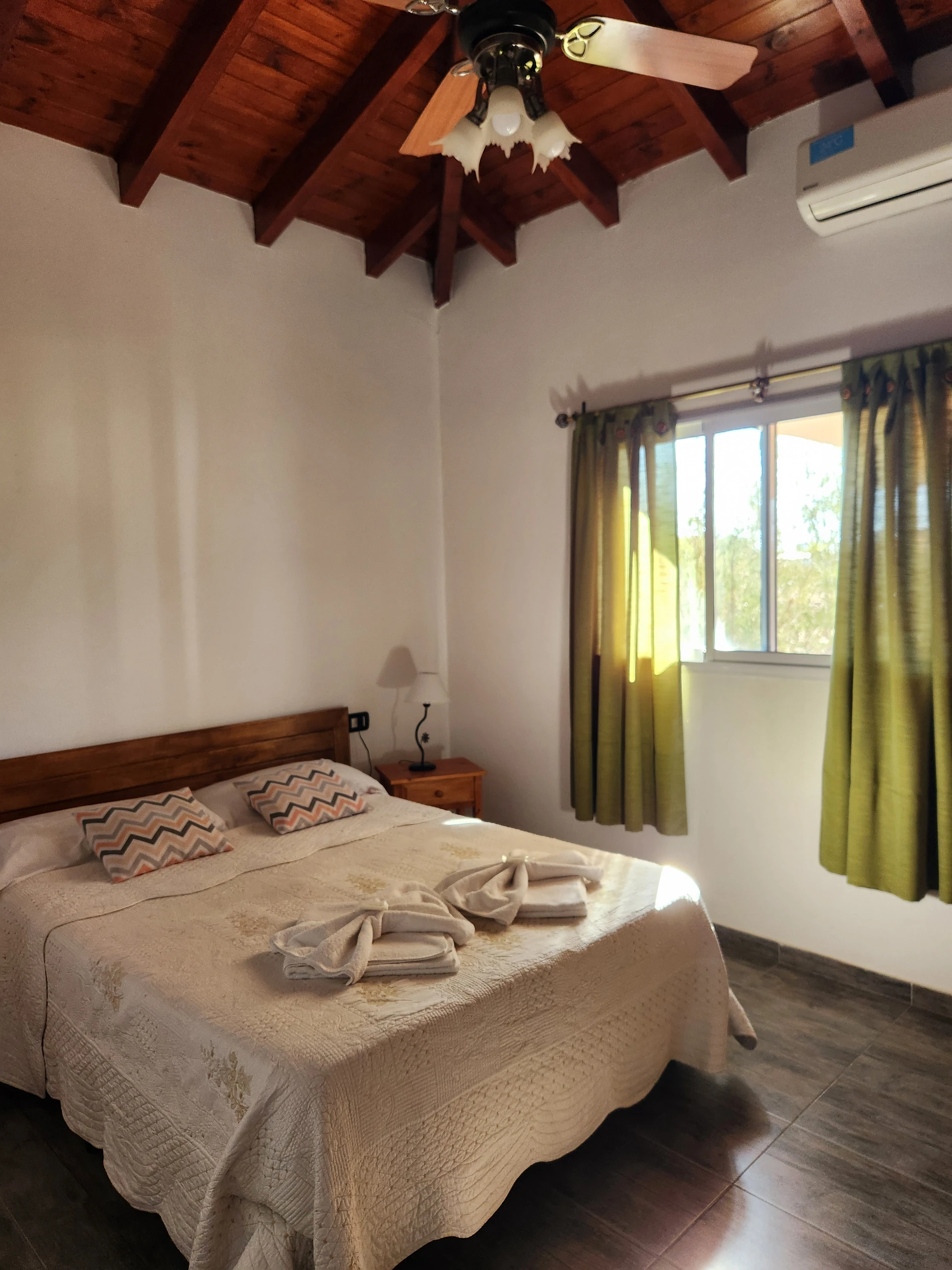Cama matrimonial junto a cortinas verdes, bajo techo de madera con ventilador.