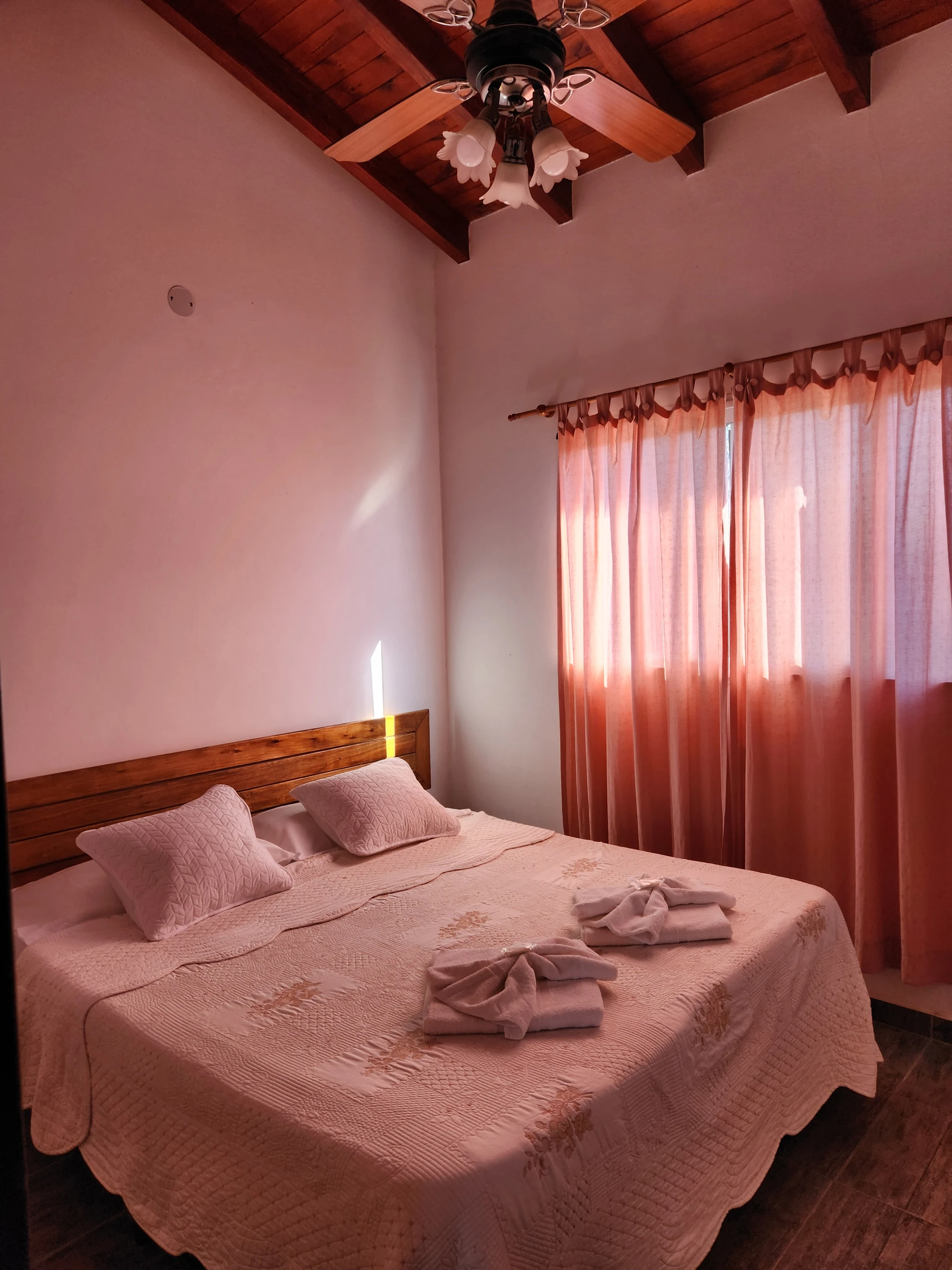 Cama matrimonial con toallas y ventana con cortinas rosadas bajo un ventilador.