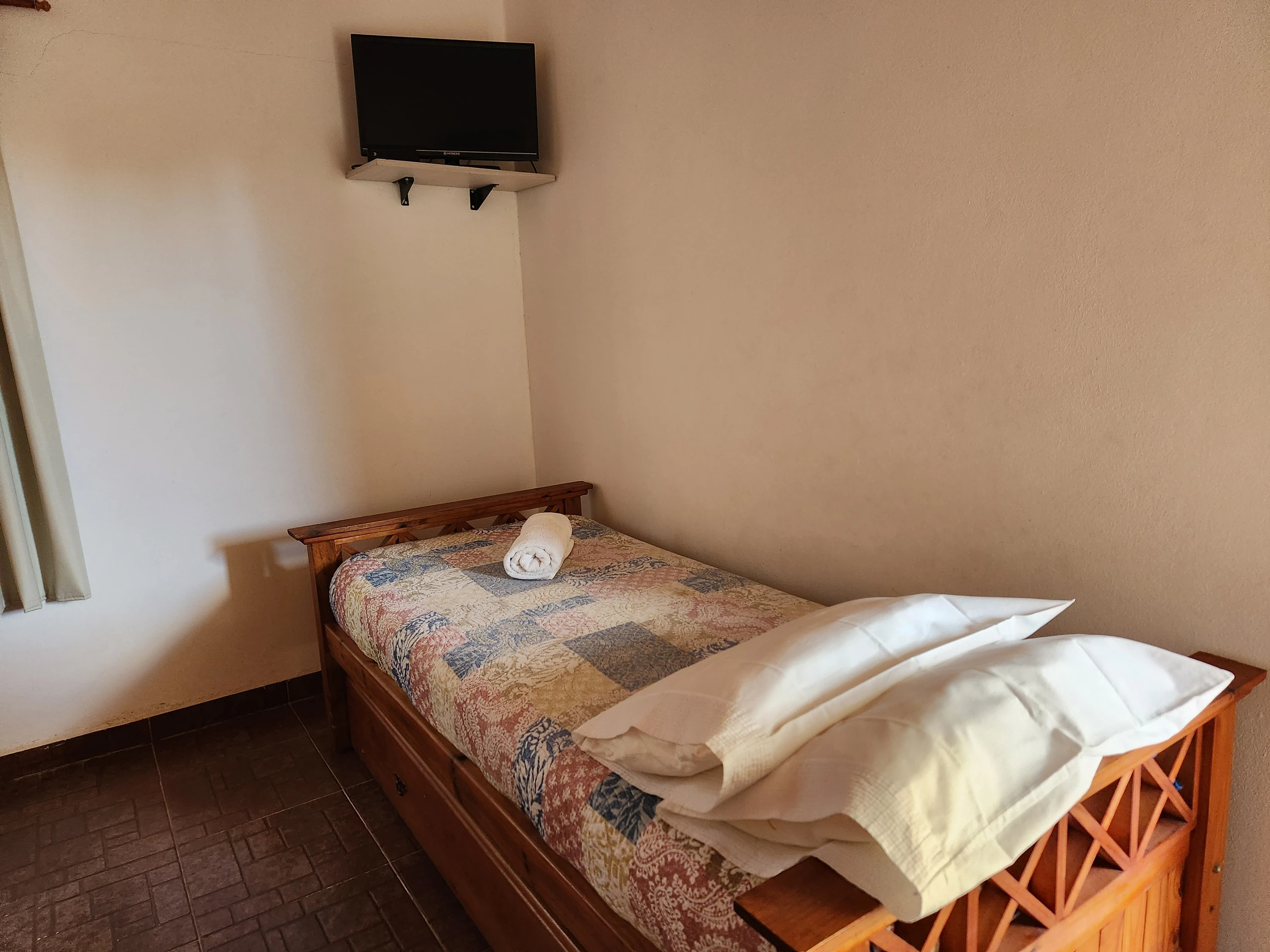 Cama individual con almohada y toallas bajo un televisor de pared.